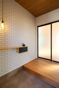 白のレンガ調の壁が特徴の玄関ホール。木製の天井と照明が、温かみのある雰囲気を演出している。【小野田産業～designers house project～】