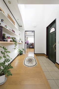 明るく広々とした玄関ホール。左側には棚があり、植物や装飾品が並べられている。右側の玄関ドアは重厚感があり、全体の雰囲気を引き締めている。【アヴィエスホーム】