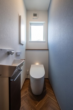 ブルーのアクセント壁とヘリンボーン柄の床が上品に調和したトイレ空間。コンパクトながらも落ち着けるデザインに仕上がっています。【イデキョウホーム】