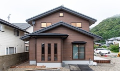左右対称の切妻屋根が印象的な、均等の取れた佇まい。木目調の外壁は落ち着いたミディアムブラウンカラー。【イシカワ】