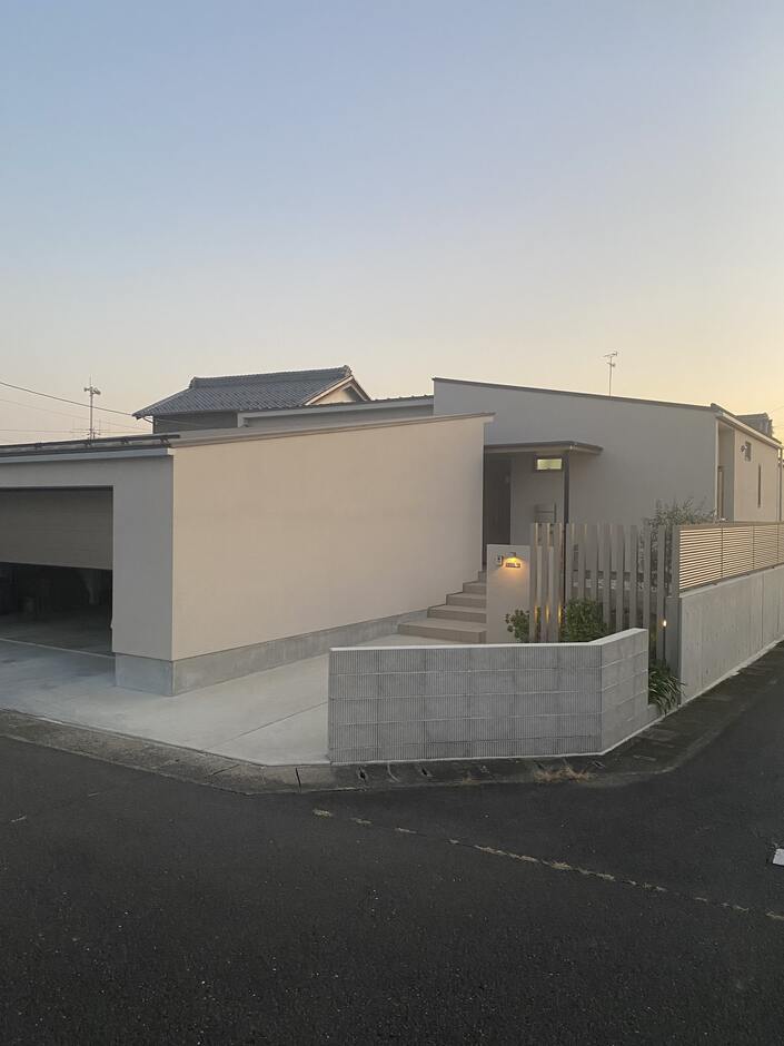 シンプルでモダンなデザインの平屋外観、ガレージシャッターを動かしている様子。柔らかな夕暮れに照らされるシックな色合いと、プライバシーを重視した構造が特徴である。