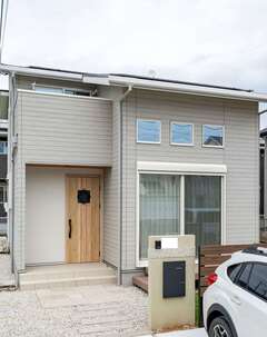 グレージュを基調とした外観。招き屋根に10kWのソーラーパネルを搭載し、低燃費な暮らしを実現。玄関ドアは迷った結果、外壁の色に合わせて明るめの木製ドアを採用した【AQURA HOME（アキュラホーム）】