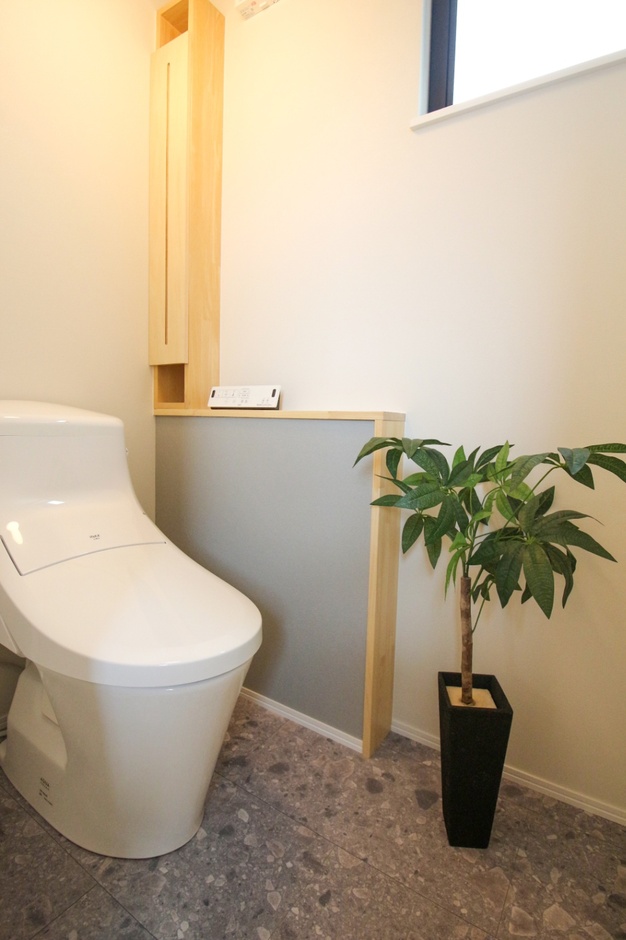 明るく清潔感あふれる内装に、実用的な造作のトイレットペーパーホルダーと、観葉植物が映えるシンプルなカウンターを配した2階のトイレ。手入れのしやすいスタイリッシュな床材を選び、限られたスペースを機能的かつおしゃれに仕上げた。