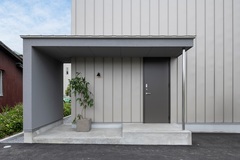 グレーの塗り壁とガルバリウムを使用した同色異素材の外観。 広くとったL字の玄関ポーチは屋根があり、 自転車やバイクの置き場に最適。【グランハウス一級建築士事務所】