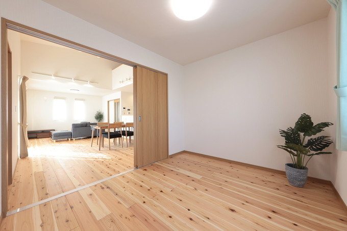 木製フローリングが広がる明るい居室。大きな窓から自然光が差し込み、開放感を演出している。隣接するリビングが見渡せるため、家族の気配を感じながら過ごせる空間。