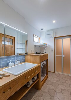 ウォークスルークローゼットからもリビングからも行き来できる洗面脱衣室。深底の医療用シンク、ブルーのサブウェイタイル、ワイドな鏡、乾太くん専用の造作棚など、奥さまのこだわりを感じる。使用頻度の高い空間がこれだけ広いと、朝の混雑もなくストレスフリー