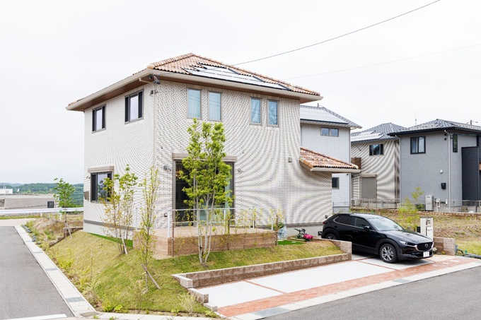 モダンなデザインの2階建て住宅の外観。庭には植樹され、自然と調和したエントランスが印象的。駐車スペースには車が停められている。