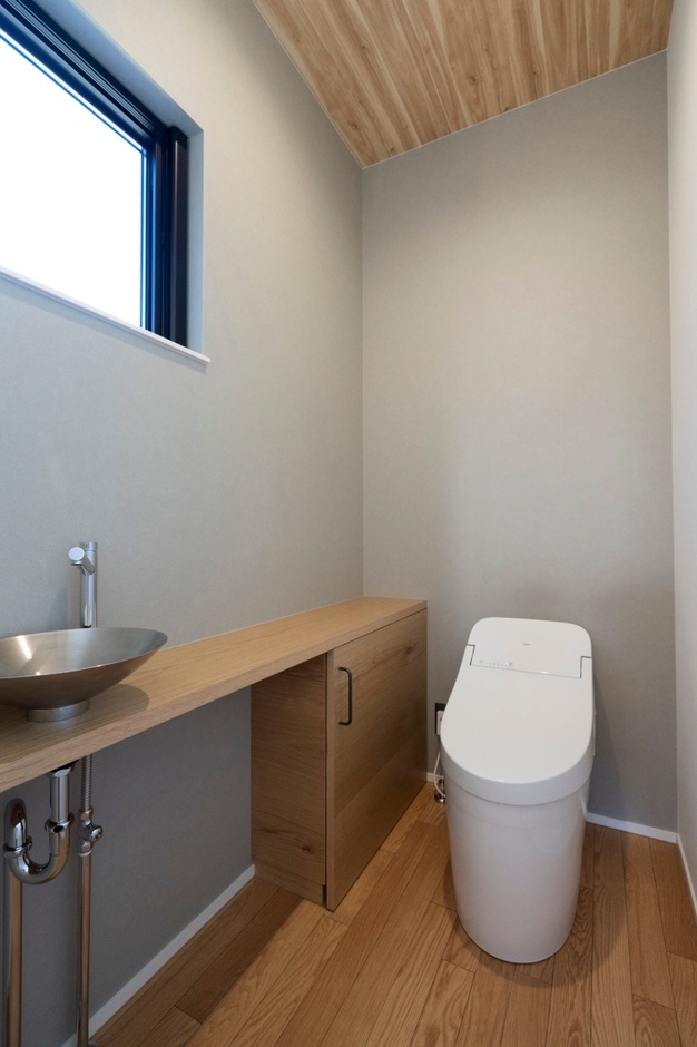 シンプルでモダンなトイレ空間。木目調の床材と天井がやわらかな印象を与え、洗練されたデザインの手洗い器と棚が機能性を高めている。