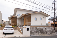 シンプルでモダンなデザインの平屋外観。立体的な屋根のラインと自然素材の木製ドアが調和し、温かみを感じさせるアプローチが特徴的である。