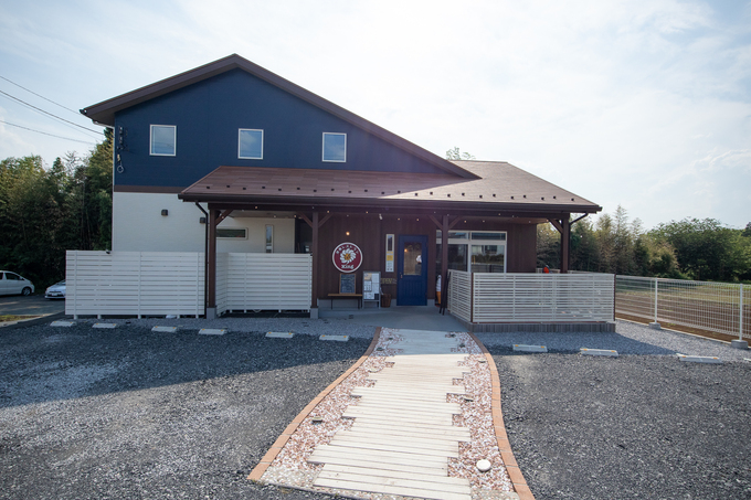 ナチュラルなウッドサイディングとモダンなブルーの組み合わせが魅力の二階建ての店舗併用住宅。広々とした玄関が特徴で、家の前には小道があり、来客を温かく迎える。